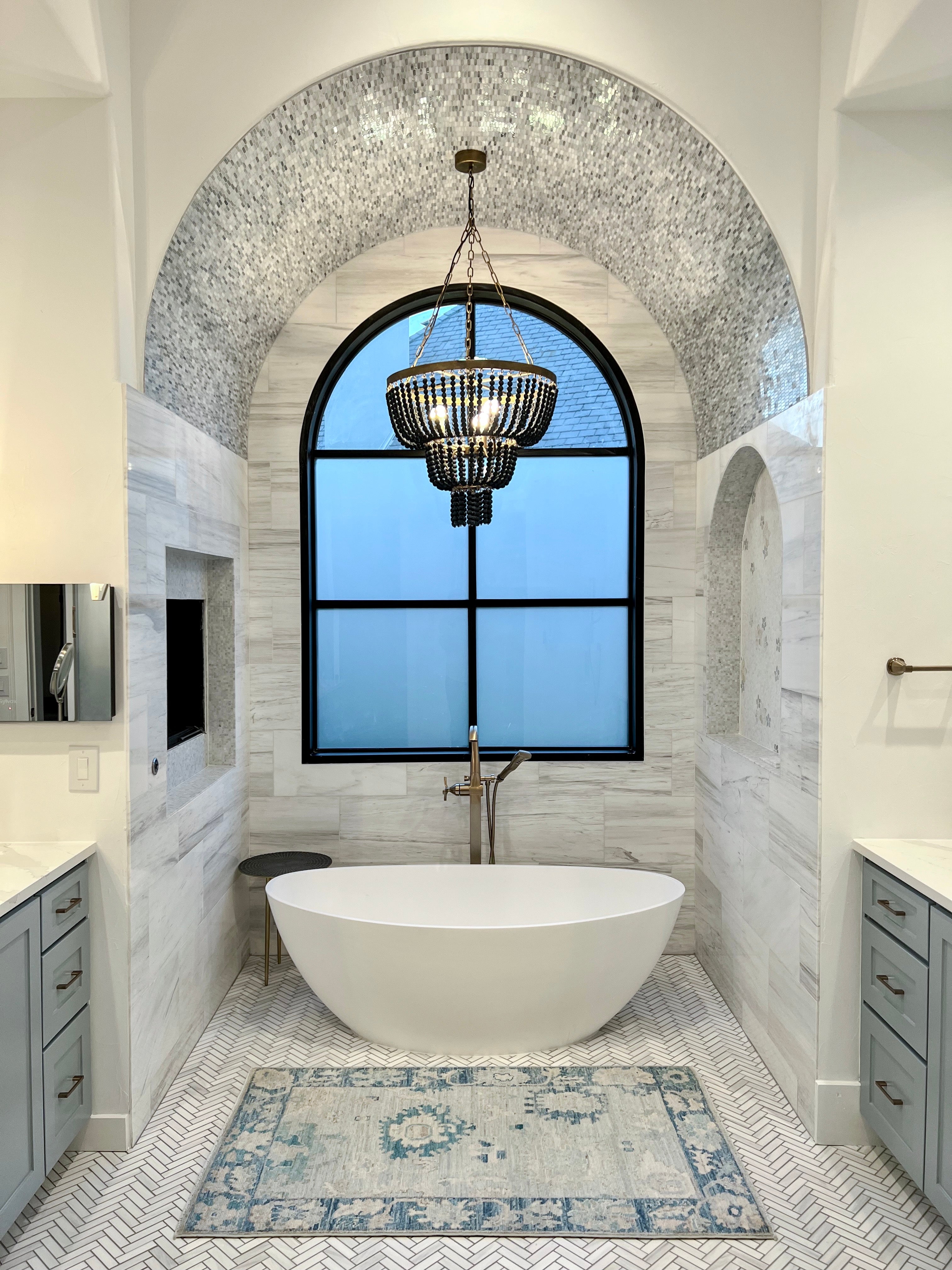 A blue and cream oushak rug from a Wooden Looms project, placed in front of a freestanding tub in a luxurious Austin, TX master bathroom with a herringbone tile floor and large arched window.
