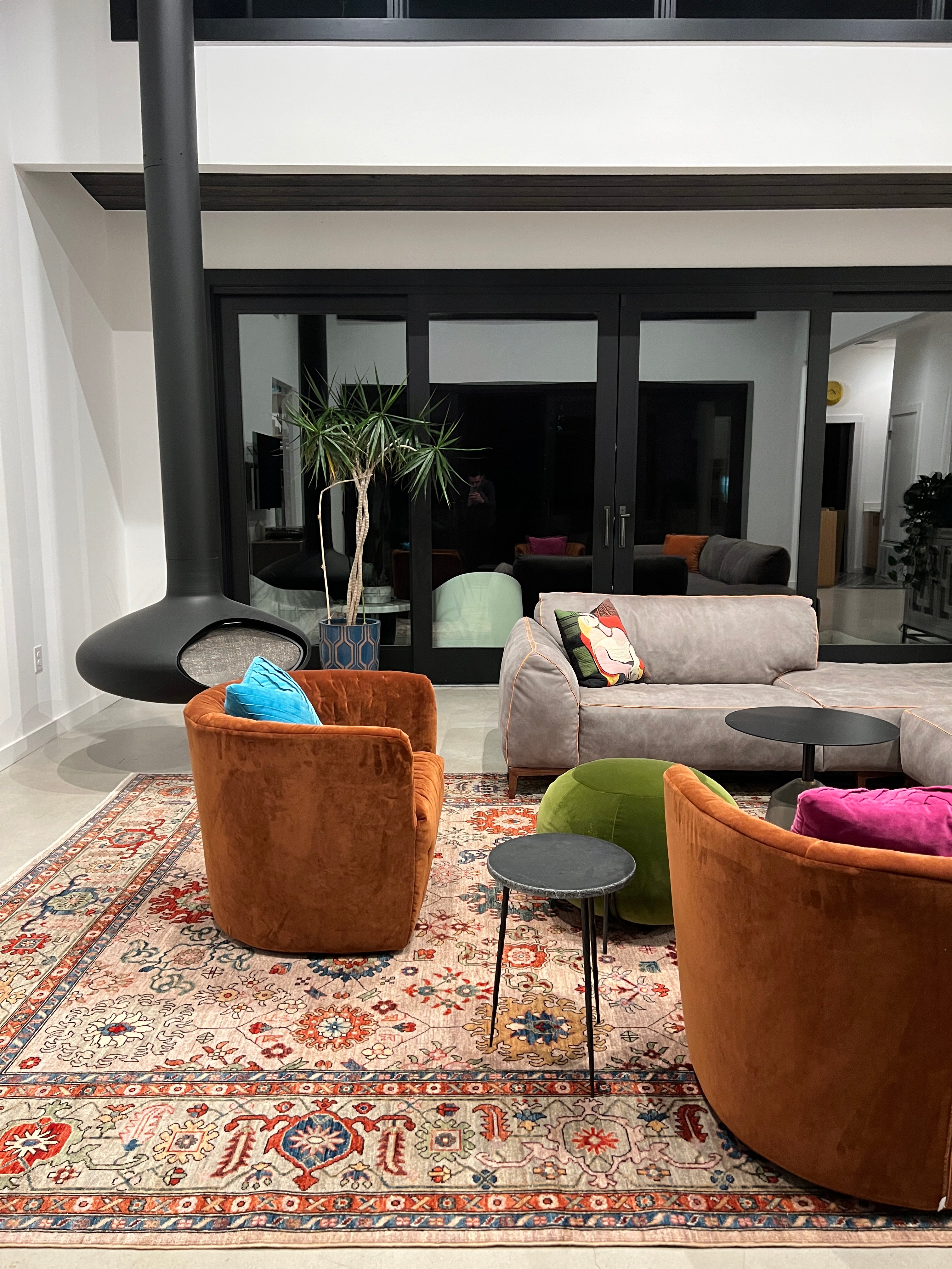 A large traditional Persian-style rug with a lilac and green pattern, featured in a Wooden Looms project in a modern Austin, TX living room with a suspended fireplace and orange velvet chairs.